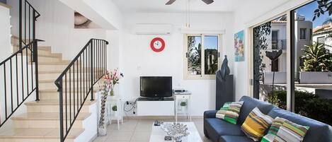 A modern living room with a staircase, a ceiling fan, a flat-screen TV, a coffee table, and a sofa with colorful cushions.