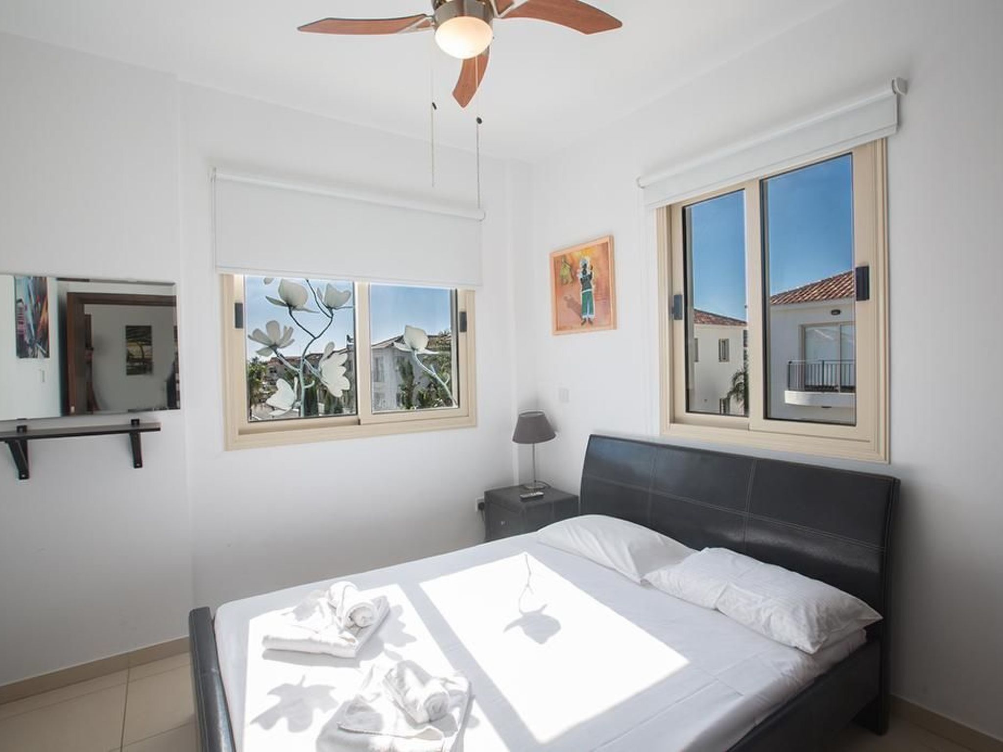 Un dormitorio con cama, ventilador de techo, ventana con vistas, mesita de noche y espejo.