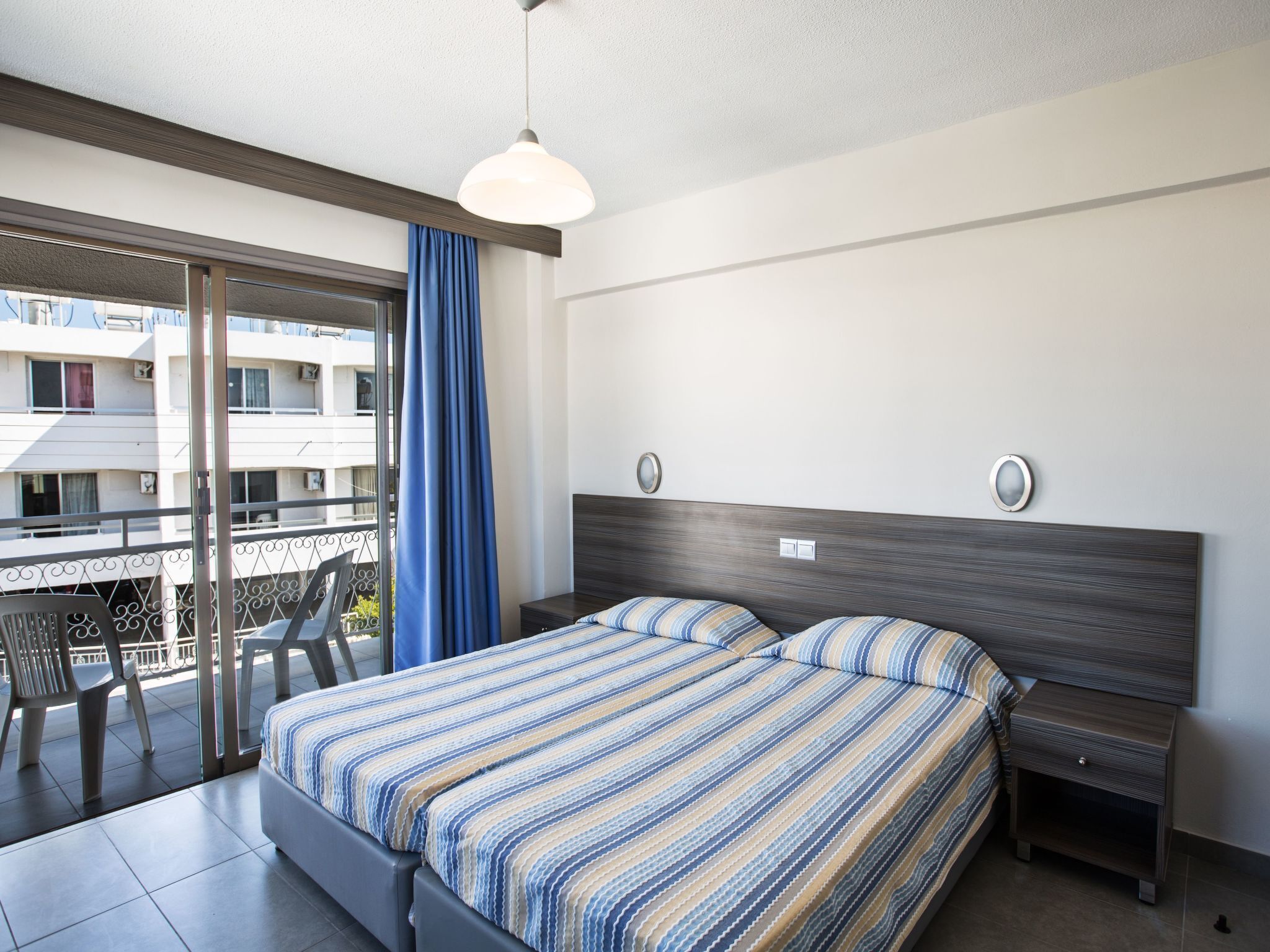 A hotel room with a bed, a nightstand, a window with curtains, and a balcony with chairs.