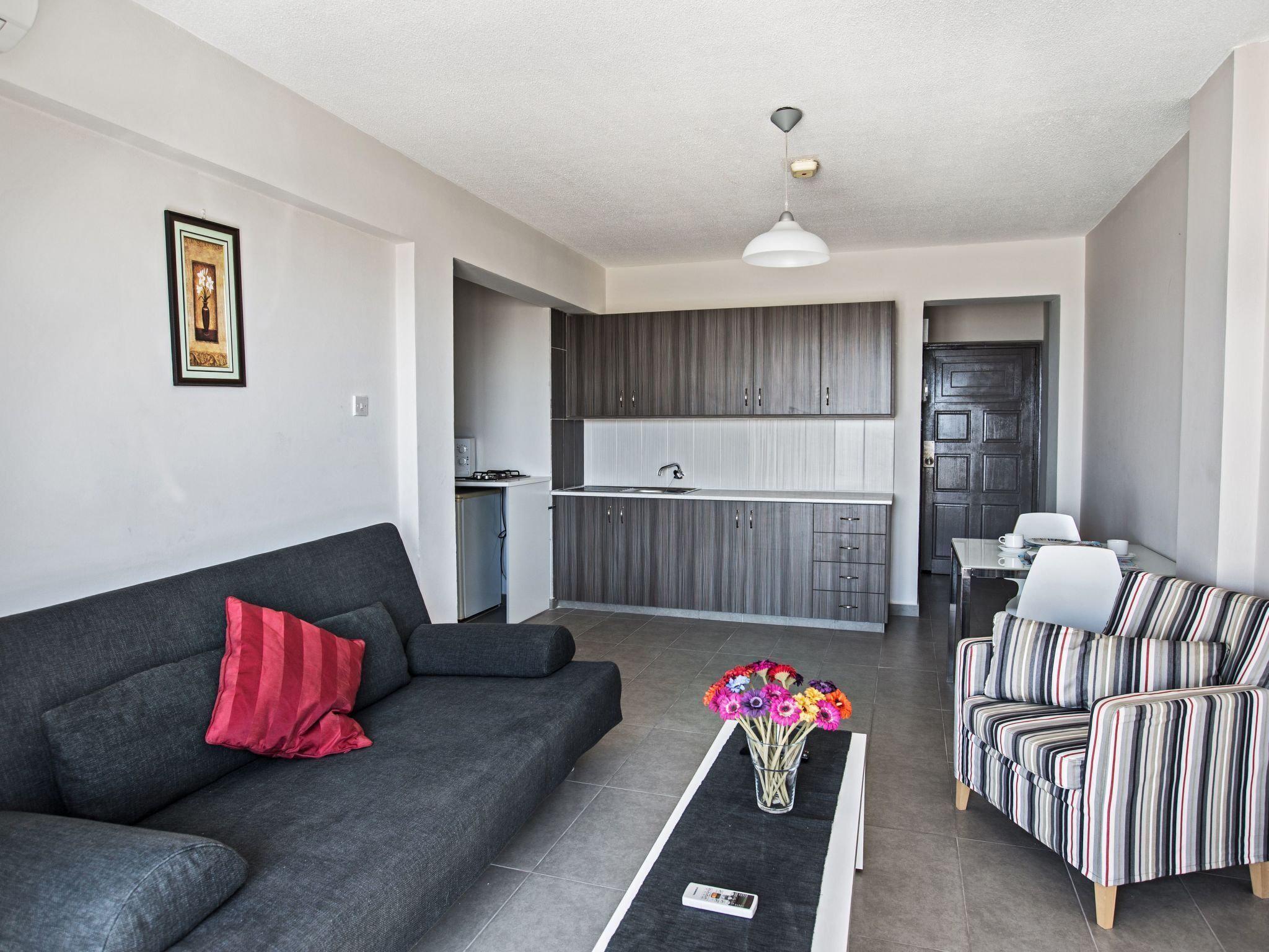 Een moderne woonkamer met een grijze bank, een gestreepte bank, een bijzettafeltje met een bloevas, en een keuken op de achtergrond.