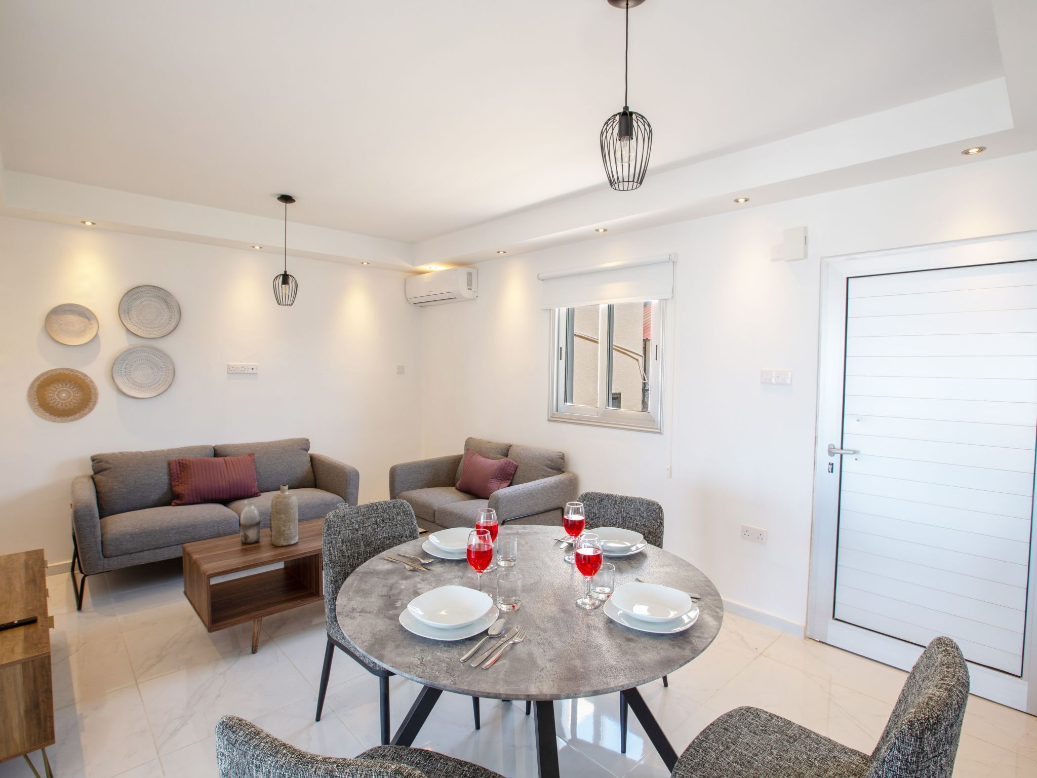 Un salon moderne avec une table à manger ronde pour quatre personnes, des canapés gris, une table basse et des assiettes décoratives accrochées au mur.