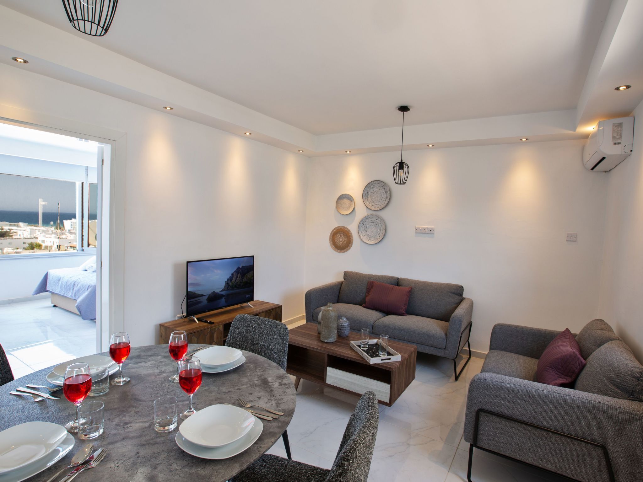 A modern living room with a dining area, a sofa, and a TV. There is a balcony with a view of the sea.