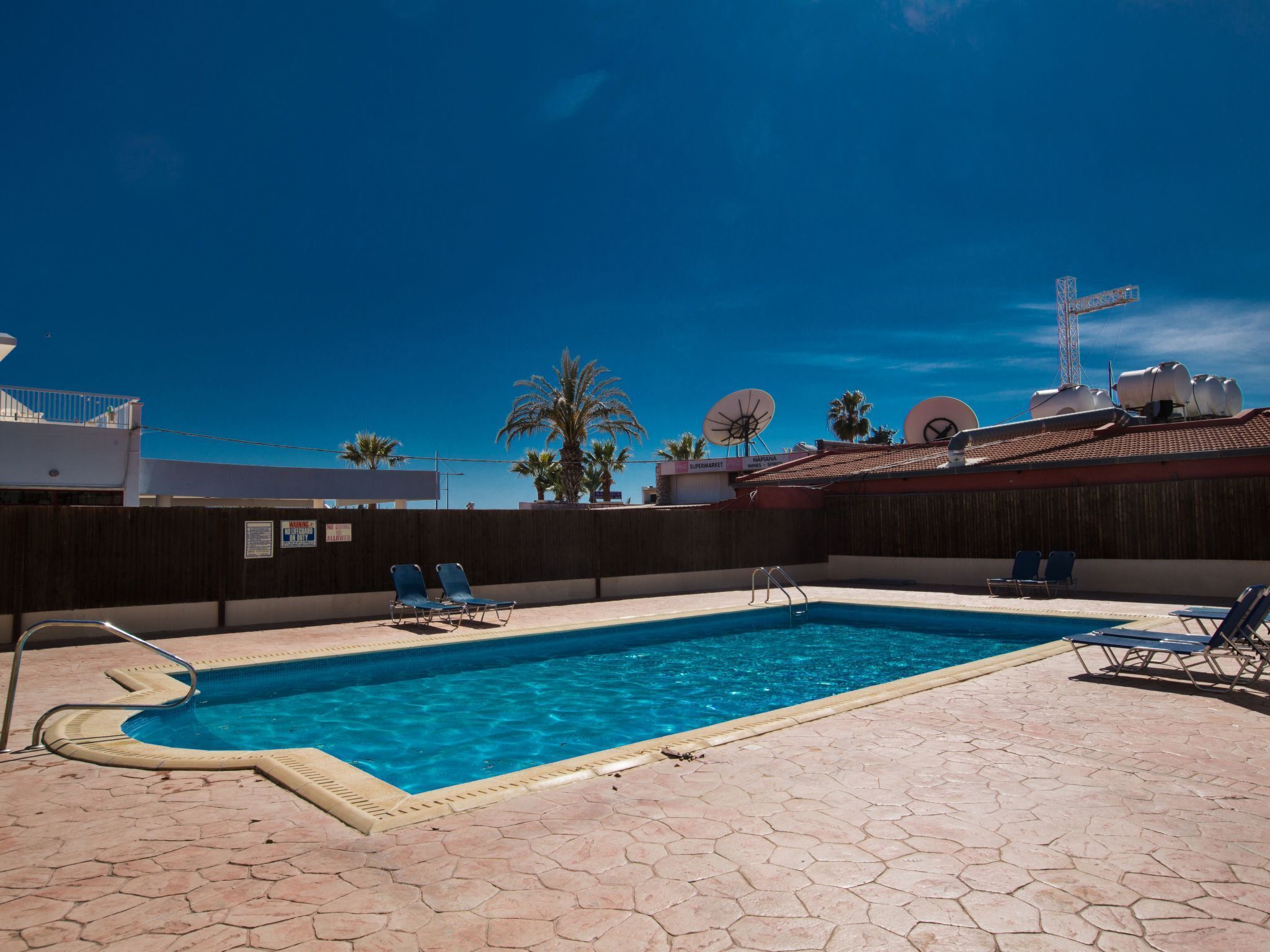 A swimming pool with lounge chairs, a wooden fence, and satellite dishes on a building.