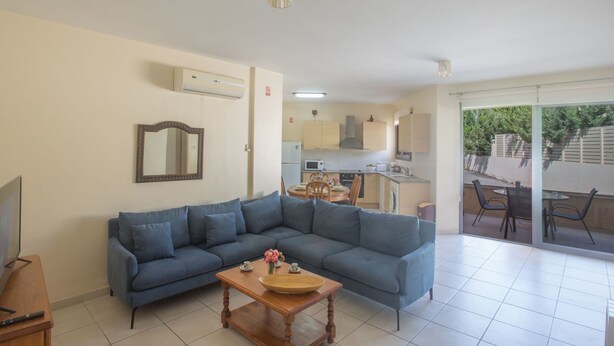 A living room with a blue sofa, a wooden coffee table, and a TV stand. There is a kitchen in the background with cabinets and a dining area. - Moderne Erdgeschosswohnung in Ayia Napa by Interhome (Ayia Napa)