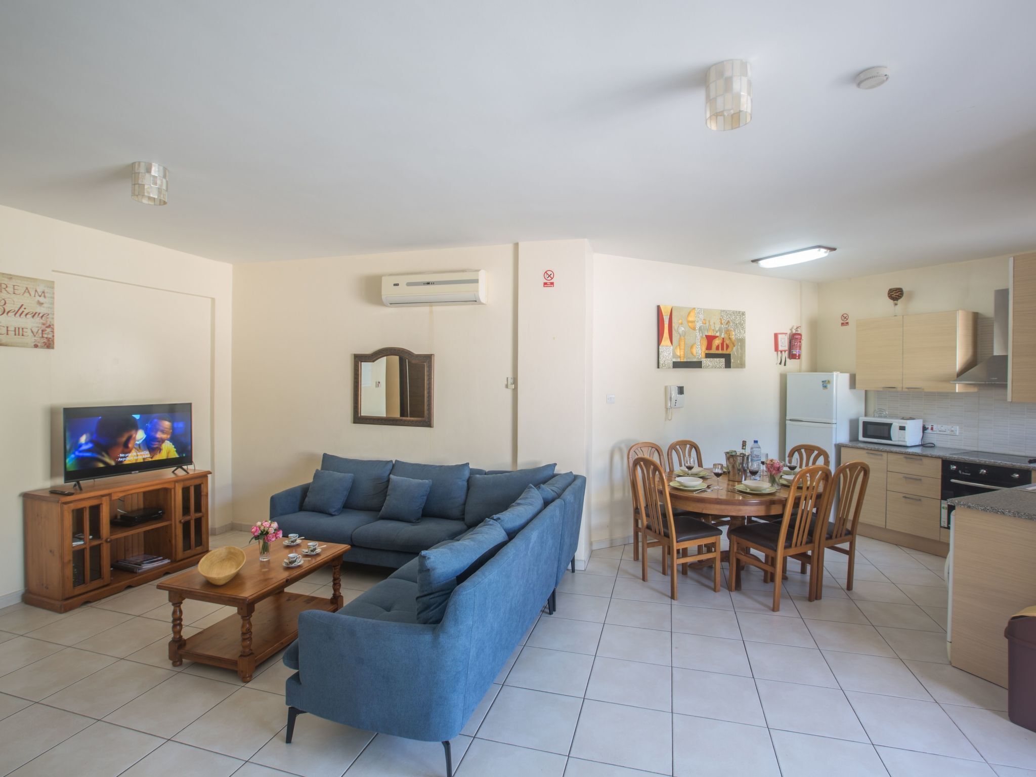 A living room with a blue sofa, a wooden coffee table, and a television. There is a dining area with a table and chairs, and a kitchen with cabinets and appliances.