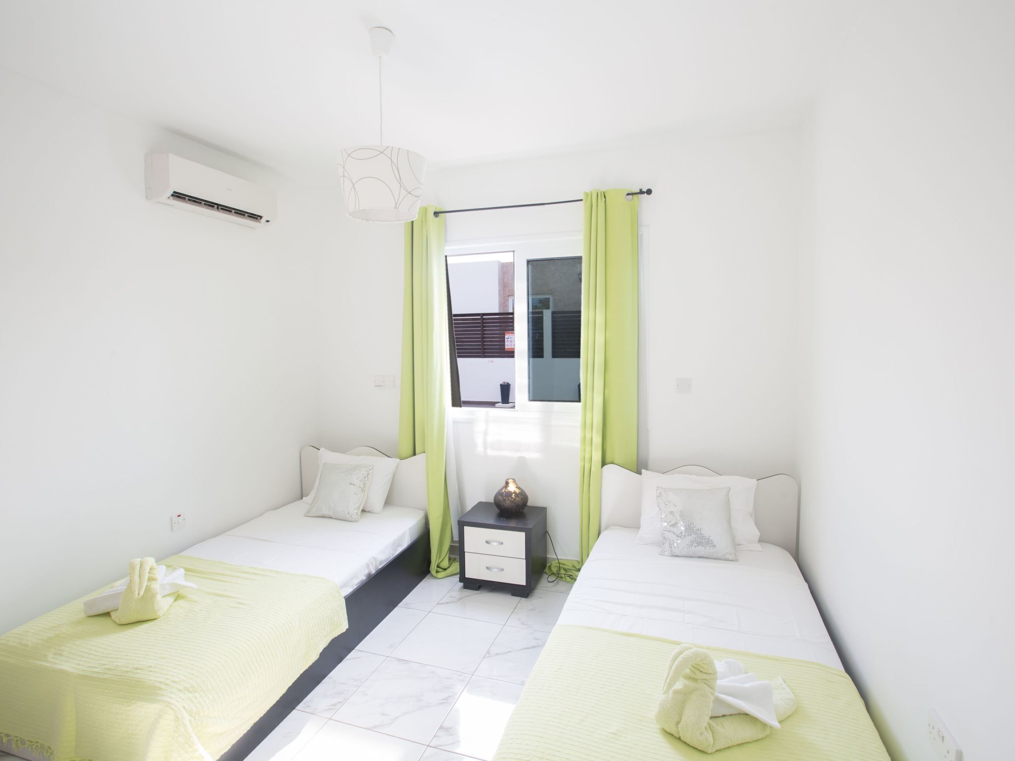 Une chambre d’hôtel avec deux lits simples, une petite table de chevet avec une lampe, une fenêtre avec des rideaux verts et un appareil de climatisation blanc.