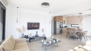 A modern living room with a sofa, coffee table, and TV. There's a kitchen with a dining area in the background.