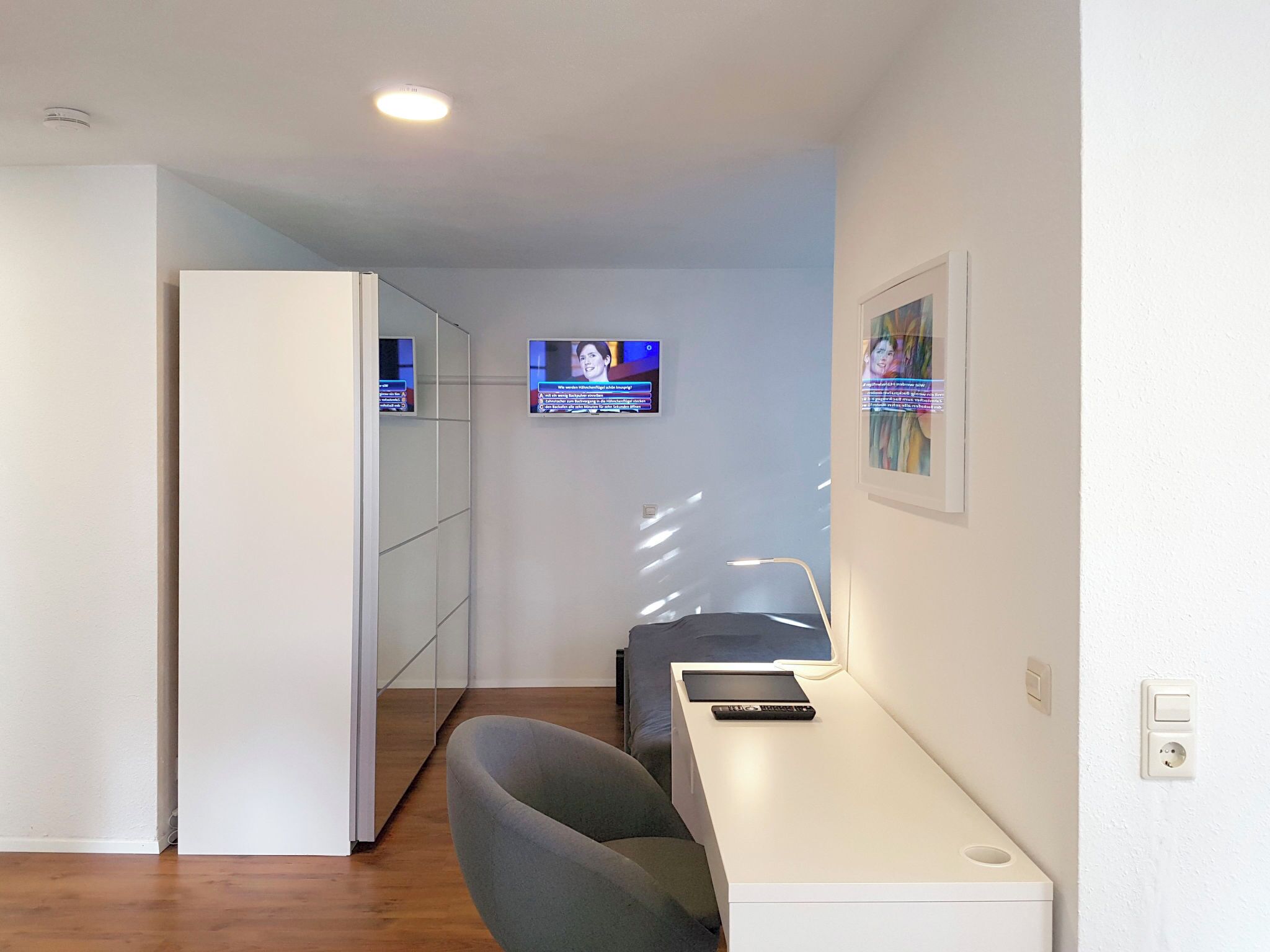 Una camera d'albergo compatta con angolo cottura, una zona salotto con una sedia e un tavolino, una televisione a parete e un quadro appeso.