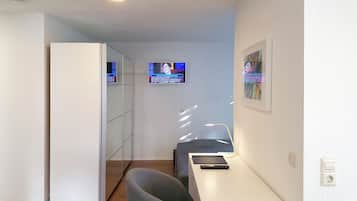 Una camera d'albergo compatta con angolo cottura, una zona salotto con una sedia e un tavolino, una televisione a parete e un quadro appeso.