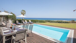 Un espace aménagé au bord de la piscine, avec une table dressée pour les repas, des chaises longues et des parasols, offrant une vue sur une mer d’un bleu limpide.