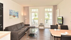 Een moderne woonkamer met een hoekbank, een glazen salontafel, een eettafel met stoelen, een televisie en grote ramen voorzien van gordijnen.