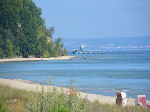 Een strand met een steiger die in het water uitloopt, omgeven door begroeiing en een verafgelegen stadsgezicht.