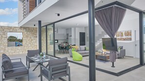 An open-plan living area with a dining table, chairs, and a sofa. There is a large window and a view of the sky. - Protaras Elite Perle Villa EP3C by Interhome (Protaras)