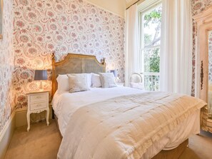 Ein Schlafzimmer mit floraler Wandgestaltung, einem hölzernen Kopfende, einer weißen Bettdecke und einem Nachttisch mit Lampe.