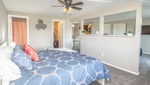 A bedroom with a bed, a ceiling fan, a mirror, and a view into the kitchen.