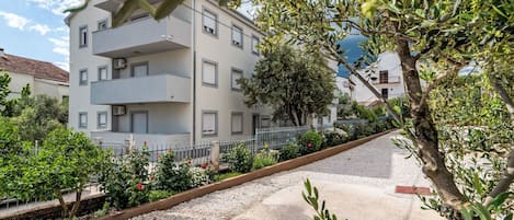 A multi-story residential building with balconies, a gravel pathway, and surrounding greenery.