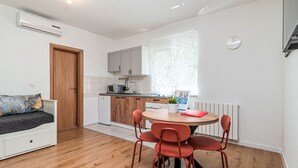 Ett kompakt bostadsutrymme med ett litet kök, en matplats med ett runt bord och röda stolar, en liten soffa och ett trägolv.