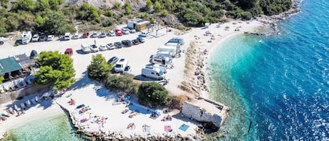 An aerial view of a coastal area with clear turquoise waters, a sandy beach, and a row of RVs parked along the shore.