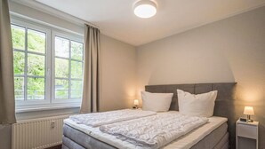 A bedroom with a double bed, bedside lamps, a window with curtains, a radiator, and a clock on the nightstand. - Bernstein by Interhome (Binz)