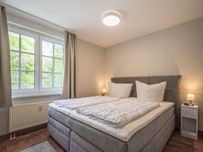 A bedroom with a double bed, bedside lamps, a window with curtains, a radiator, and a clock on the nightstand. - Bernstein by Interhome (Binz)