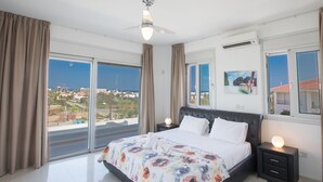 Quarto com uma cama grande, um ventilador de teto e vista da cidade através de janelas amplas.