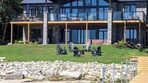 A large, modern two-story house with a stone facade, large windows, and a deck overlooking a body of water. - Experience Ultimate Luxury at our Exquisite Lakehouse. Large family gatherings! (Erinsville)