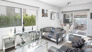 A modern living room with a sofa, coffee table, and a glass-top table with decorative items. There is a dining area with chairs and a table, and a large window providing a view of greenery outside. - Moderne 4-Schlafzimmer-Villa im Zentrum von Protaras by Interhome (Protaras)