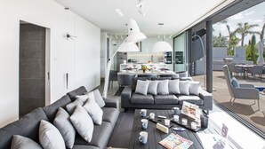 A modern living room with a grey sectional sofa, a coffee table with magazines and cups, and a large window offering a view of the outdoor patio area.
