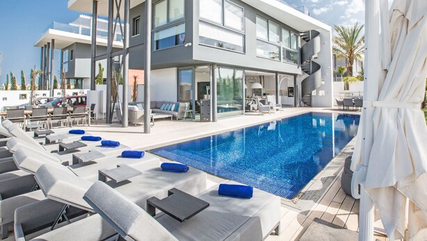 A modern two-story hotel with a large outdoor swimming pool, lounge chairs, and a covered seating area.