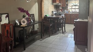 A dining area with a tiled floor, wooden chairs, a table with a vase of flowers, and a kitchen in the background with wooden cabinets and a window. - Charming 4-bedroom house perfect for your getaway. Panoramic view from balcony (Guadalupe)