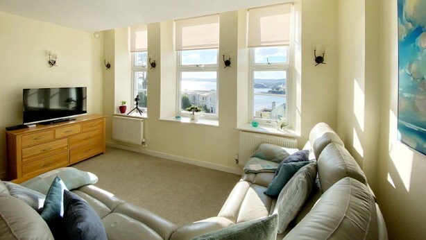 A living room with a sofa, a TV, and a view of the city through large windows. - Wohnung mit Meerblick in Torquay by Interhome (Torquay)