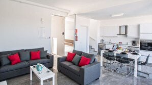 Um apartamento moderno com dois sofás cinzentos com almofadas vermelhas, uma mesa de centro branca, uma área de jantar com mesa de vidro e cadeiras de metal, e uma cozinha com armários e eletrodomésticos brancos.