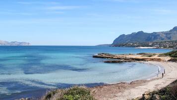 A coastal beach with clear waters, rocky outcrops, and a distant mountain range.