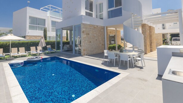 A modern poolside area with a clear blue pool, white outdoor furniture, and a multi-level building with glass doors and a spiral staircase. - Luxuriöse Villa Vie Bleu by Interhome (Protaras)