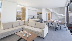 A modern living room with a sectional sofa, a coffee table with cups and flowers, a dining area with chairs and a table set for a meal, and a kitchen in the background. - Ferienvilla Vie Bleu mit großem Pool by Interhome (Protaras)