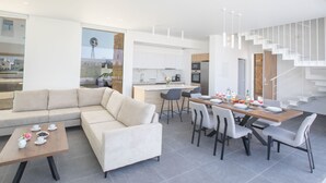 A modern living room with a sofa and coffee table, a dining area with chairs and a table set for two, and a kitchen with white cabinets and a built-in oven. - Ferienvilla Vie Bleu mit großem Pool by Interhome (Protaras)