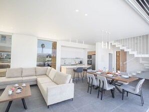 Ein modernes Wohnzimmer mit Sofa und Couchtisch, ein Essbereich mit Stühlen und Tisch für zwei Personen sowie eine Küche mit weißen Schränken und einem Einbauofen.