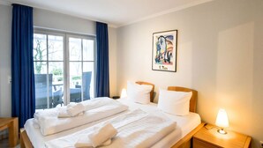 A hotel room with a large bed, a bedside table with a lamp, a window with blue curtains, and a framed artwork on the wall. - Seeschloss Binz Apartment 26, 2 Minuten zum Strand by Interhome (Binz)