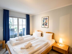 Ein Hotelzimmer mit einem großen Bett, einem Nachttisch mit Lampe, einem Fenster mit blauen Vorhängen und einem gerahmten Kunstwerk an der Wand.