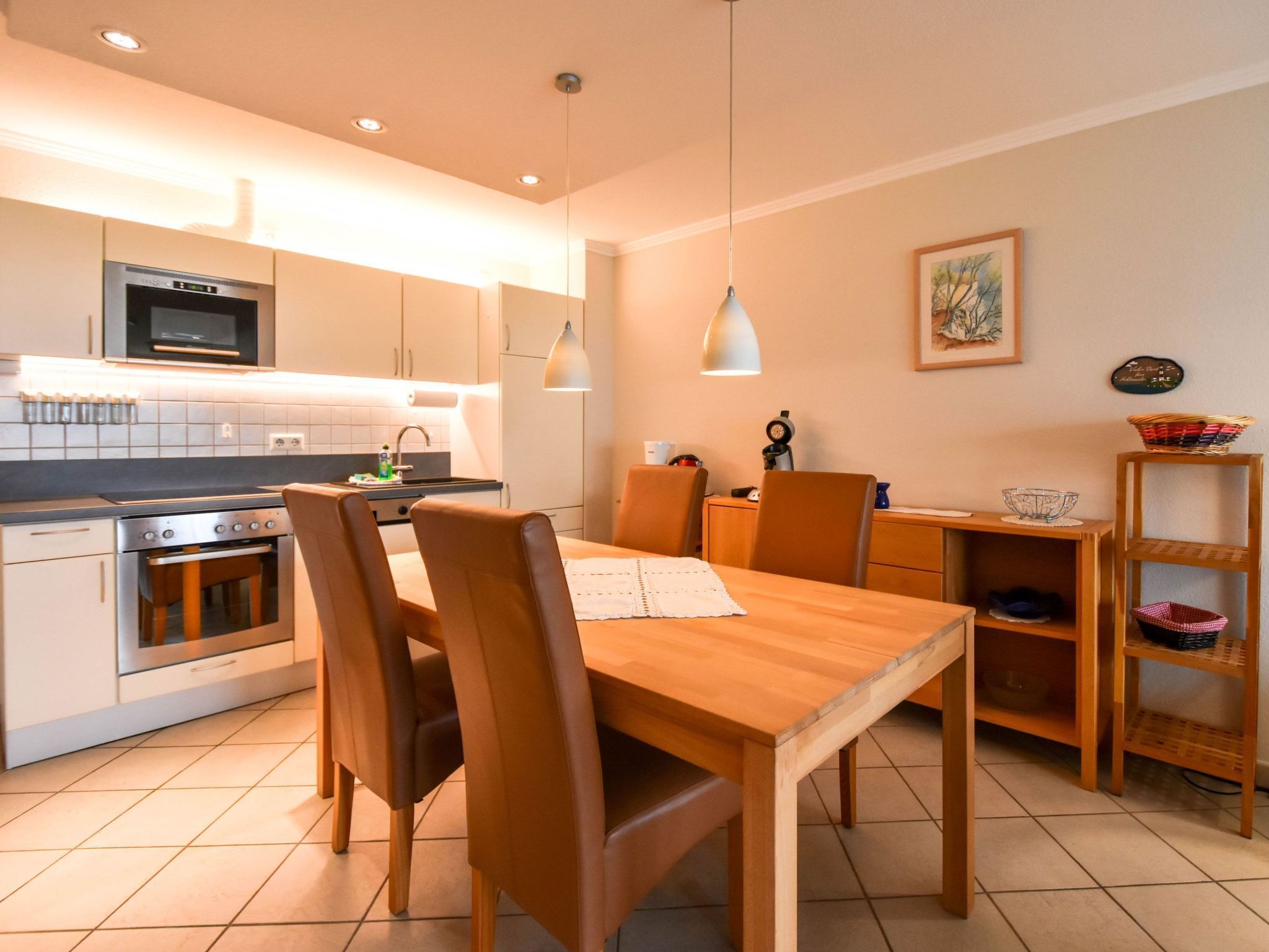 A modern kitchen with a dining area, featuring stainless steel appliances, wooden furniture, and pendant lighting.