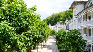 A tree-lined street with white buildings featuring balconies and a central pathway. - Appartement NeunUndZwanzig in Ostseebad Binz by Interhome (Binz)