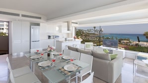A modern kitchen with white cabinets and a dining area with a glass table set for four. A large window offers a view of the ocean and palm trees. - Luxuriöse Wohnung am Strand von Thalassa by Interhome (Paralimni)
