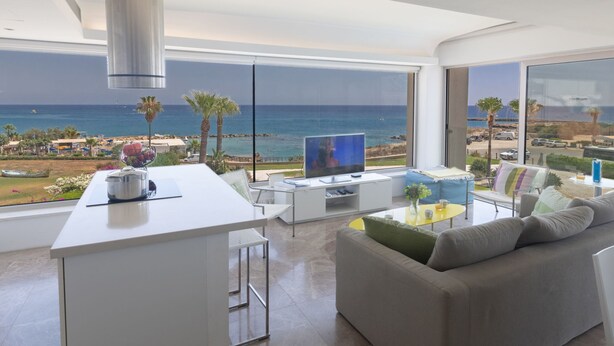 A modern living room with a large window overlooking a beach, a kitchen area, and a seating area with a sofa and coffee table. - Luxuriöse Wohnung am Strand von Thalassa by Interhome (Paralimni)