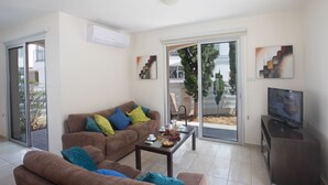 Un soggiorno moderno con un divano marrone, un tavolino da caffè e una televisione. Una porta scorrevole in vetro dà accesso a una zona esterna con un albero e una panchina.