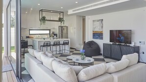 Una cocina moderna con una isla central, un comedor con sofá y sillas, y una sala de estar con televisor y sillón de peluche.