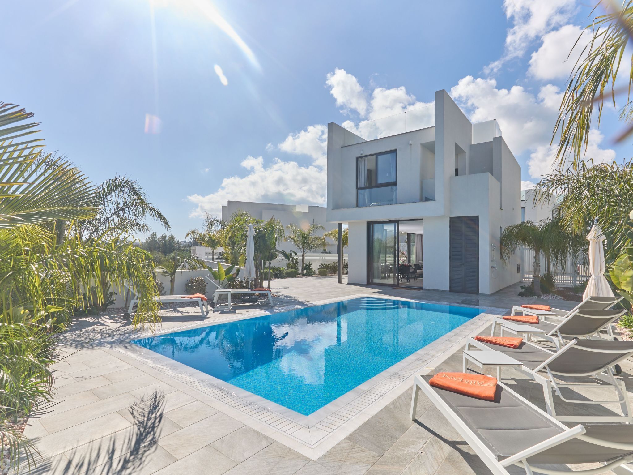 A modern two-story house with a swimming pool, surrounded by palm trees and lounge chairs.