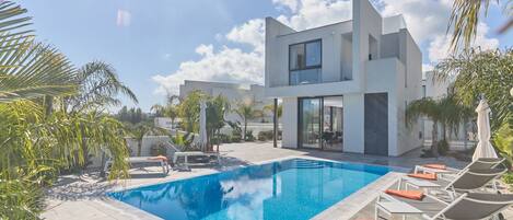 A modern two-story house with a swimming pool, surrounded by palm trees and lounge chairs.