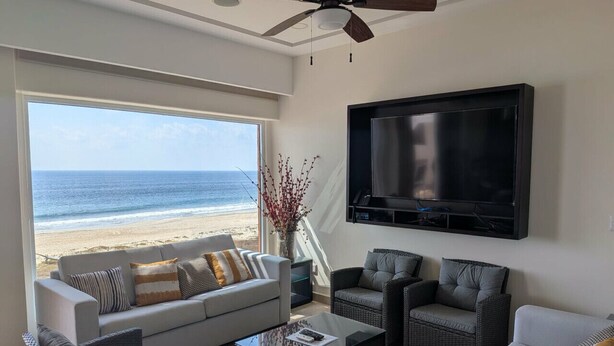 Een woonkamer met een groot raam dat uitkijkt op een strand, een flatscreen televisie aan de muur en een plafondventilator.