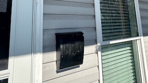 Ein Haus mit einer weißen Tür, einem Fenster mit Rollläden, einem schwarzen Briefkasten und einer wandmontierten Leuchte.