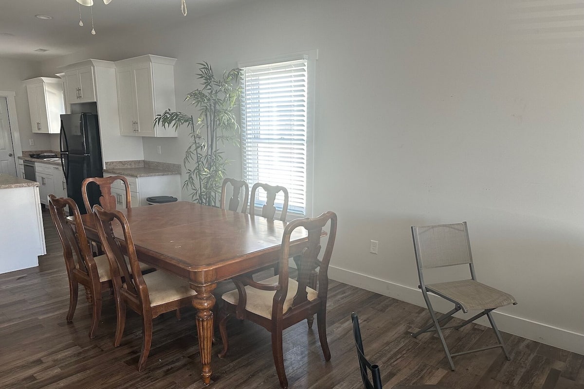 Un comedor con mesa y sillas de madera, una cocina con armarios blancos y una ventana con persianas.
